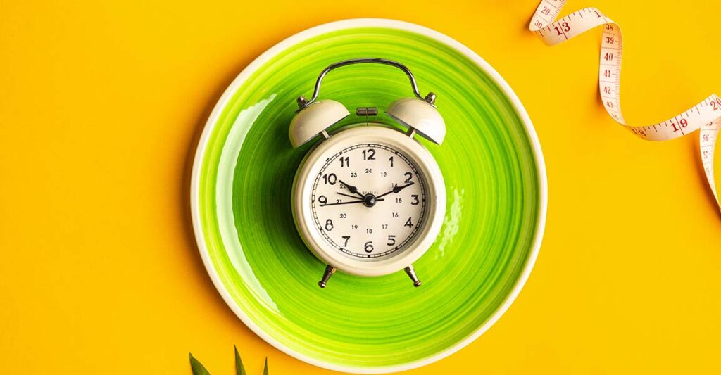 Fotografía de un reloj sobre un plato y un vibrante fondo amarillo, con una cinta métrica en el lado derecho de la imagen, que representa el ayuno intermitente.