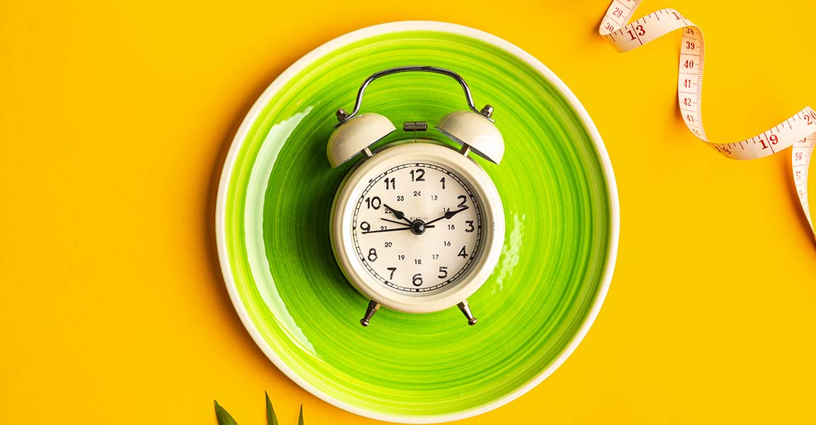 Fotografía de un reloj sobre un plato y un vibrante fondo amarillo, con una cinta métrica en el lado derecho de la imagen, que representa el ayuno intermitente.